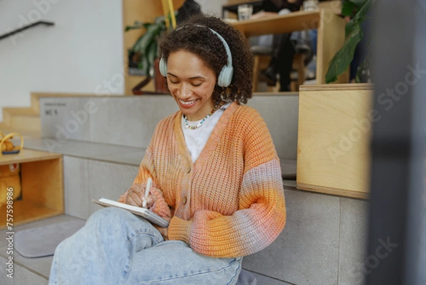 Fototapeta a woman wearing headphones is sitting on the floor writing in a notebook