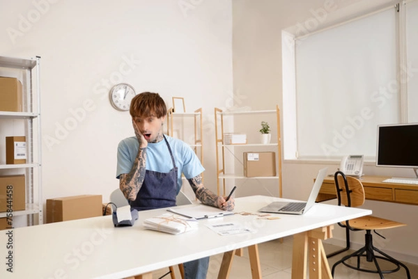 Fototapeta Shocked online store seller working with payment terminal at table in warehouse