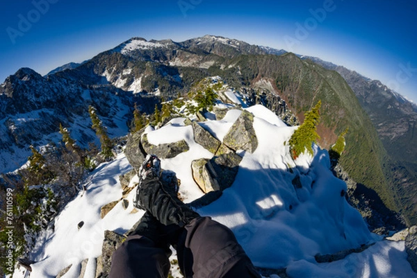Fototapeta Adventurous athletic male hiker, sitting in a snowy boulder field with his legs outstretched before jagged snowy mountains on a beautiful sunny day.