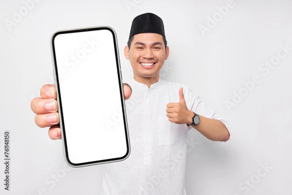 Fototapeta Smiling young Asian Muslim man showing display mobile phone screen, showing thumb up gesture isolated on white background. People religious Islam lifestyle concept. celebration Ramadan and ied Mubarak