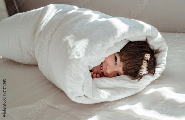 Fototapeta Little happy toddler boy wrapped in blanket like a roll looking at camera smiling having fun on the bed.
