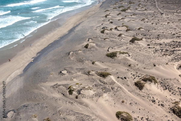 Fototapeta Aerial view of sandy dunes with scarce vegetation and a wavy ocean with few surfers on the Famara beach, Lanzarote, Canary Islands
