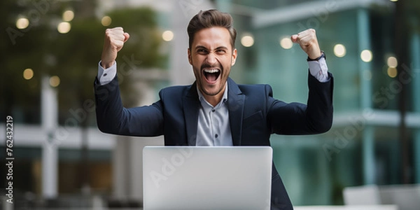 Obraz A young businessman celebrating success with arms raised in front of a laptop, looking a laptop in a happy and successful pose