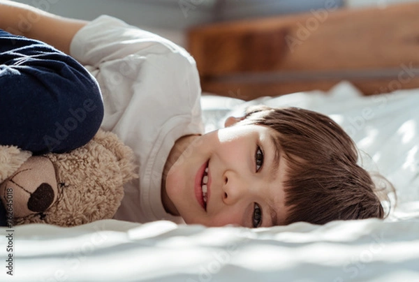 Fototapeta Portrait of little happy toddler boy lying on bed with his teddy bear plush toy looking at camera smiling at sunny morning
