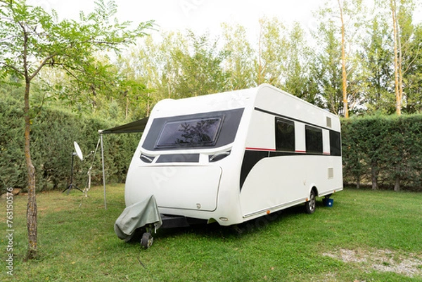 Fototapeta A white trailer,a motorhome for travel,a motorhome,camper,trailer without a car against a background of beautiful bushes stands on short green lawn grass.Technologies of transport, means of travel