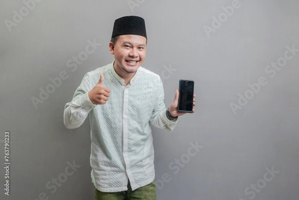 Fototapeta Portrait of an Asian Muslim man smiling with showing thumbs up and his smartphone