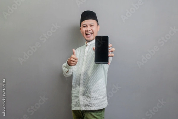 Fototapeta Portrait of an Asian Muslim man smiling with showing thumbs up and his smartphone