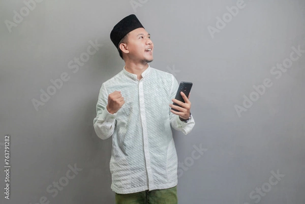 Fototapeta Asian Muslim man clenching his fist and holding his smartphone while looking at an empty space