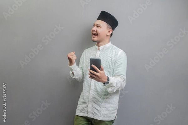 Fototapeta Asian Muslim man clenching his fist and holding his smartphone while looking at an empty space