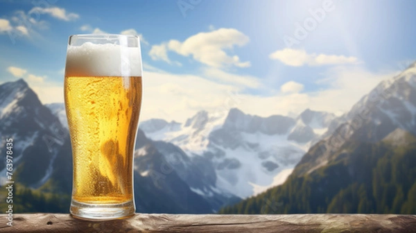 Fototapeta Glass of lager beer on a wooden table with snowy mountain peaks background.