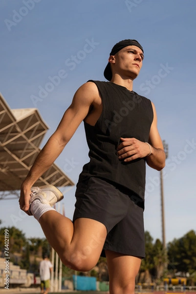 Obraz Young male athlete warming up his legs and stretching. Preparing for competition in a sports facility. Vertical photo
