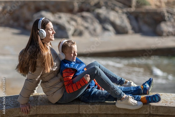 Obraz  A mother and her son are sitting with a smiling face, on a beach listening to music with headphones