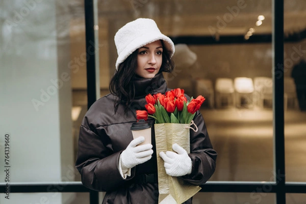 Obraz Attractive brunette woman walking near glass office building in moody cloudy windy weather with bouquet of red tulips flowers, drink coffee tea hot beverage. International Women`s Day 8th March concep