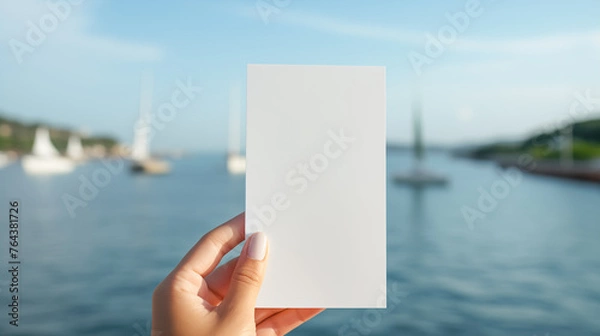 Fototapeta Female hand holding a piece of white paper on a blurred background of an ocean bay with yachts. Copy space for text.