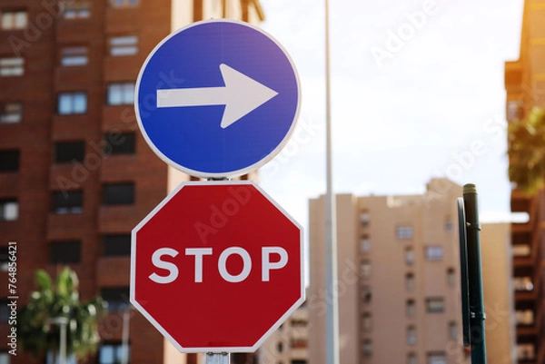 Fototapeta Traffic rules. Red road sign stop and blue and white right directional arrow on residential buildings background. Street sign direction pointing turn right, but need to stop before. Two road signs.