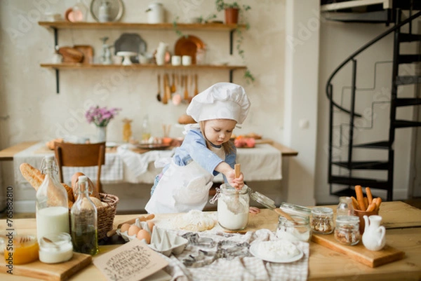 Fototapeta Girl cook 3 years old in a beautiful kitchen, in a chef's hat, in an apron, daylight