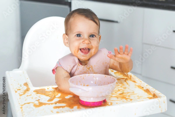 Fototapeta happy fun cheerful messy baby 9 months old sits in a highchair and eats lure vegetable puree soup, the concept of baby food and childhood