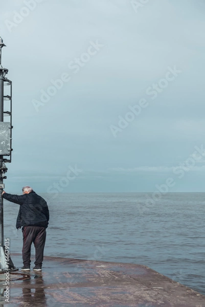 Fototapeta Senior man leaning on the headlight of the pier in a Riviera Romagnola beach location. Dramatic sky in a winter afternoon. Winter sea.