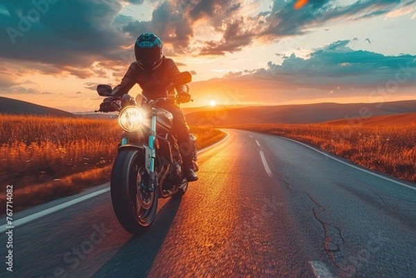 Obraz A man is riding a motorcycle down a road at sunset. The sky is filled with clouds, and the sun is setting in the distance