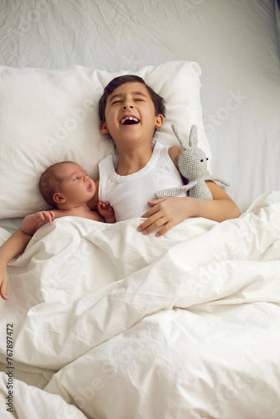 Obraz cheerful portrait of a boy of seven years old lying with his sister baby on a pillow on a white big bed with a crocheted toy hare , light from the window.