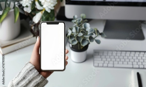 Fototapeta Mock up. A woman's hand holds a phone against the background of a workspace with a laptop and a cup of coffee