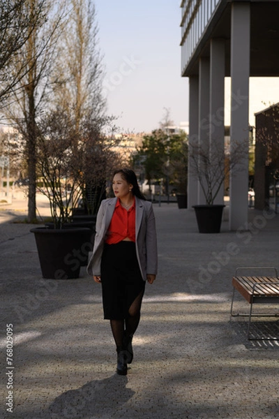 Fototapeta Confident woman in business attire walking through an urban park with modern architecture, wearing a grey blazer, red blouse, black skirt, and boots.