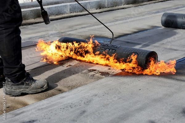 Fototapeta Professional construction workers using a blowtorch to glue modified bitumen roll sheet over a concrete road.