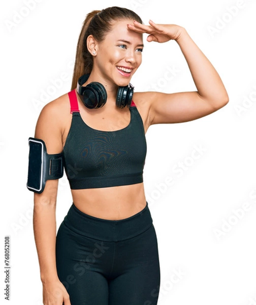 Obraz Beautiful blonde woman wearing gym clothes and using headphones very happy and smiling looking far away with hand over head. searching concept.