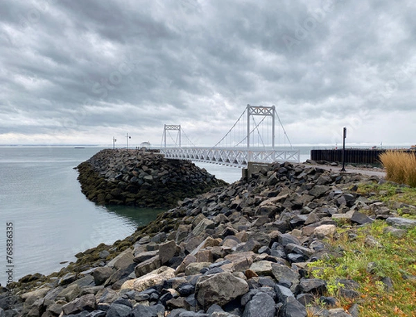 Fototapeta Pier on the coast. Structure of a pedestrian bridge on a quay. Walkway at the river's edge on a cloudy day. White gate on a rocky bank. Boat and buoy in the distance.