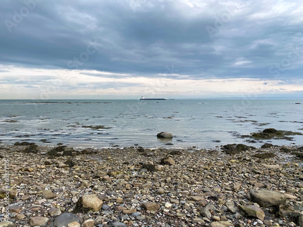 Fototapeta Beach and rocks on a cloudy day. Coastal beach at low tide. Stones on the coast. Cargo boat in the distance on the river.