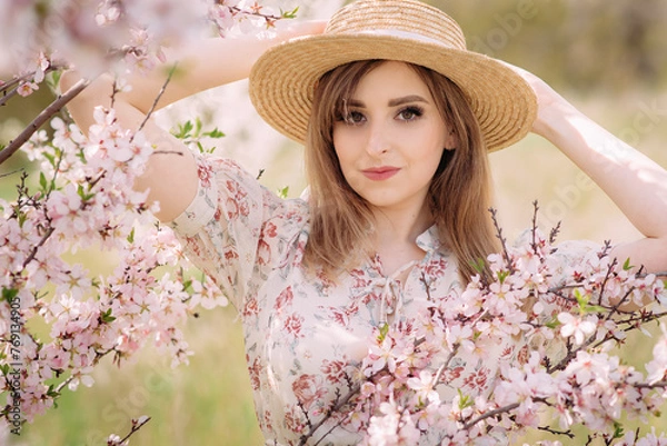Fototapeta A woman in a white dress with a colorful pattern enjoys the aroma and poses with a hat on her head. A beautiful young woman stands near a blooming spring tree in the garden. Lifestyle concept.