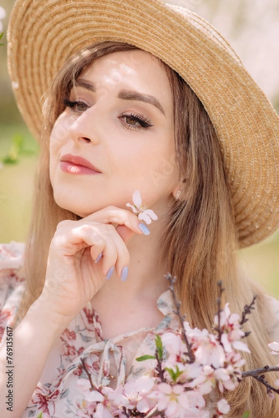Fototapeta A woman in a white dress with a colorful pattern poses with her eyes closed. A beautiful young woman in a hat is standing near a blooming spring tree in the garden and holding a flower in the garden