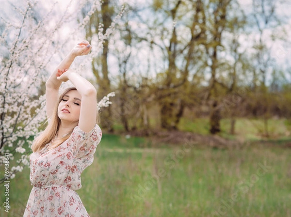 Fototapeta A woman in a white dress with a colorful pattern enjoys the aroma of flowers. A girl is standing near a blooming spring tree in the garden with her arms raised. Background with space for text