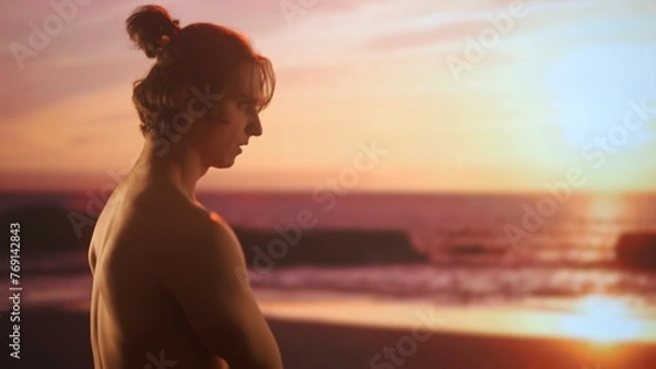 Fototapeta Profile of a young man with bare torso admiring a sunset, his face illuminated by the warm golden light of the twilight sky.