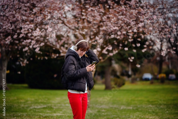 Fototapeta Engrossed in her smartphone and music, a young woman stands under the spring bloom of cherry blossoms, her headphones suggesting a moment of leisure and relaxation