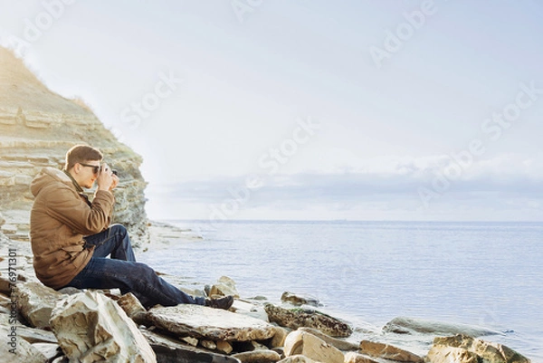 Fototapeta Traveler man sitting on coastline and taking photographs