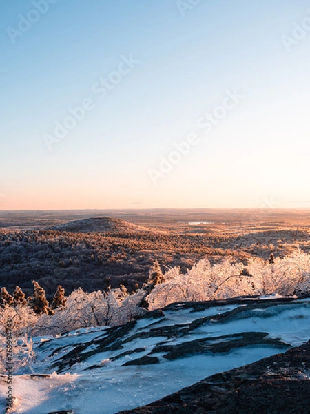 Fototapeta Golden hour above the summit of Mount Watatic in Ashburnham Massachusetts. The mountain and forest is covered in ice and snow, which is reflecting the golden sun. A small mountain and lake are visible