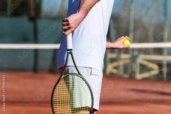 Obraz Close up of young boy with racket playing tennis on a clay court during a university tournament. The tennis player is wearing a white sports suit and is intent on serving the set point