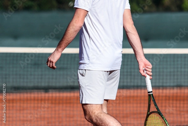 Obraz Close up of young boy with racket playing tennis on a clay court during a university tournament. The tennis player is wearing a white sports suit and is intent on serving the set point