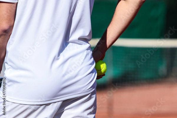 Obraz Close up of young boy with racket playing tennis on a clay court during a university tournament. The tennis player is wearing a white sports suit and is intent on serving the set point