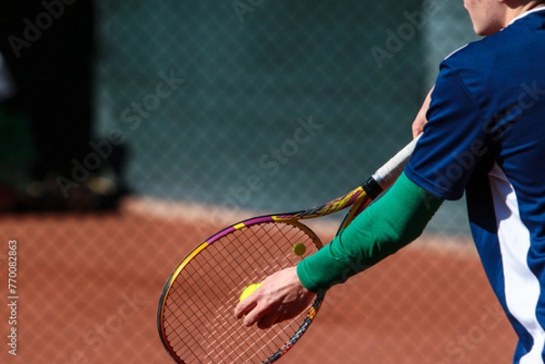 Obraz Detail of young boy with racket playing tennis on a clay court during a university tournament. the athlete is wearing a blue sports t-shirt and is preparing to serve for set point