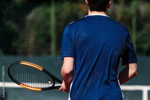 Obraz Close up of young boy with racket playing tennis on a clay court during a university tournament. the athlete is wearing a blue sports shirt.