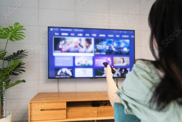 Fototapeta An Asian woman is watching TV in the living room at home, holding a TV remote control