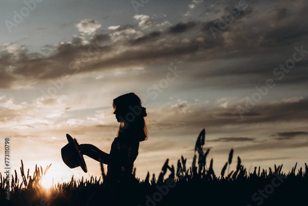 Obraz silhouette of a girl holding a hat at sunset in the field