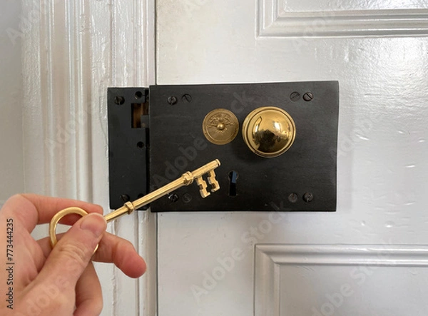 Fototapeta Antique door lock. Golden door handle with antique gold-engraved crest. Ancient lock with key. Old metal key in the keyhole. Hand holding the key.