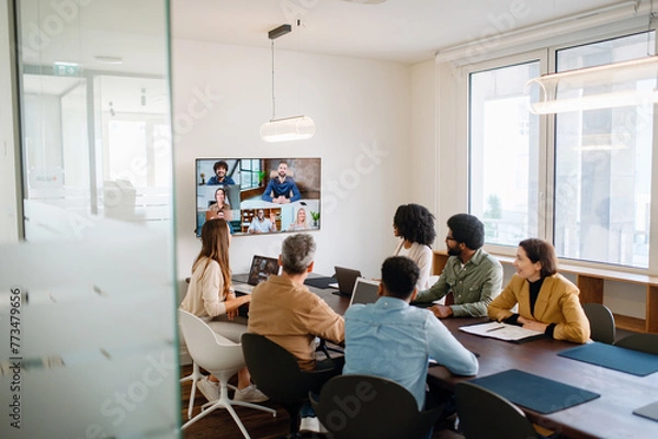 Fototapeta A team actively engages with remote colleagues via a large screen, illustrating the modern hybrid work model where technology bridges the gap between in-office and remote team members