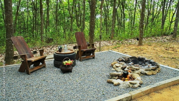 Fototapeta Two rock fire pit with wood table and chairs on gravel floor in the garden background. Stone fire pit with tire table decoration and wooden reclining chair on pebble ground.