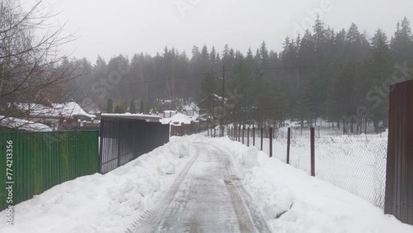 Fototapeta After the snowfall snow lies on the ground and roofs of houses. Along the street there are metal and wooden fences and electricity poles. Tire tracks can be seen on the snow-covered road. Foggy