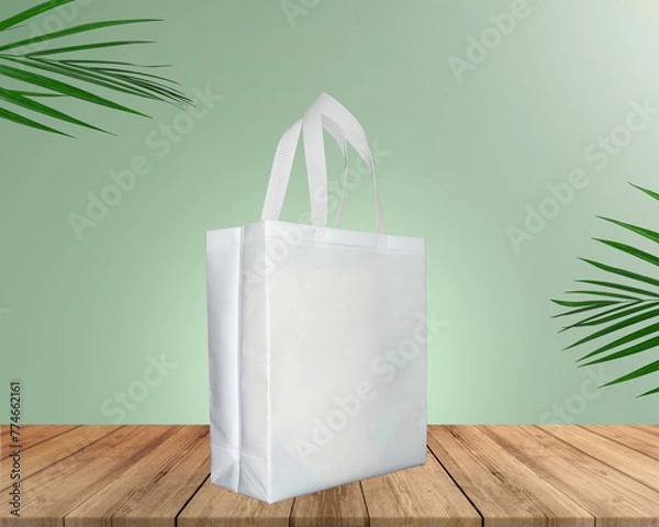 Fototapeta White Color Eco Friendly Tote Bag With Loop Strap. Non Woven Fabric Bag isolated on wood table with plants green background With copyspace for text