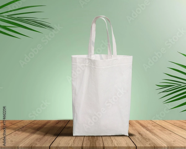 Fototapeta White Color Eco Friendly Tote Bag With Loop Strap. Non Woven Fabric Bag isolated on wood table with plants green background With copyspace for text